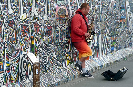 A street musician plays saxophone while leaning against a heavily graffitied wall.