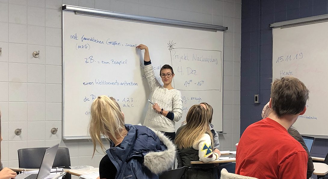 A female graduate student teaches grammar in front of a whiteboard to a mixed group of students.