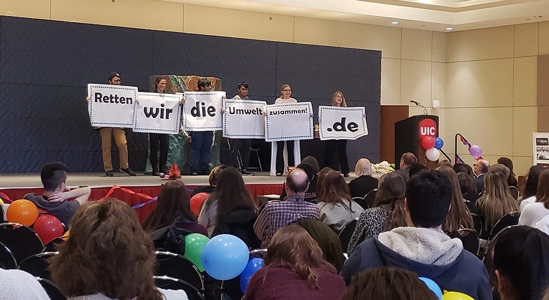 a mixed group of students stand on a stage before a mixed audience holding signs spelling out the sentence 